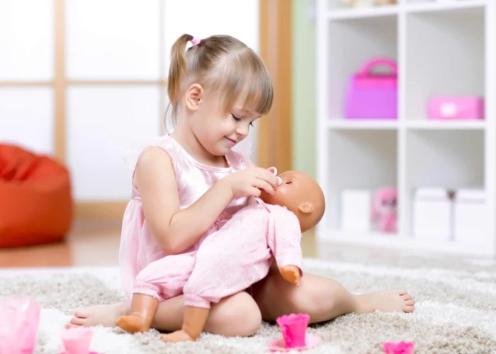 A smiling toddler girl in a pink dress plays with Cute Dolls for Toddlers, gently feeding her baby doll in a cozy, colorful playroom.