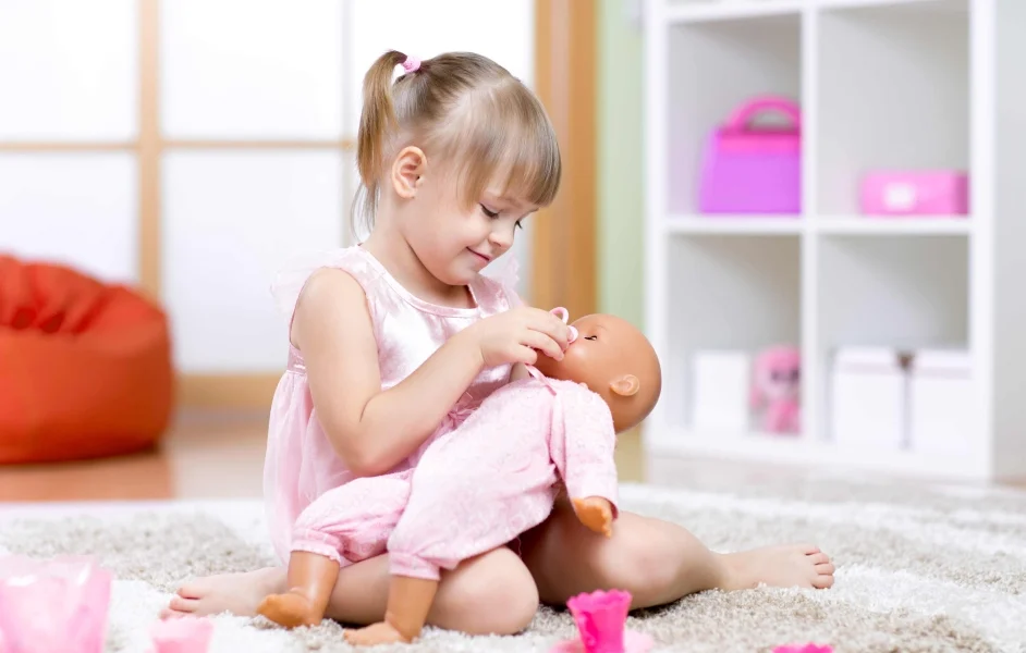 Soft, Snuggly and Sweet: The Best Cute Dolls for Toddlers A smiling toddler girl in a pink dress plays with Cute Dolls for Toddlers, gently feeding her baby doll in a cozy, colorful playroom.