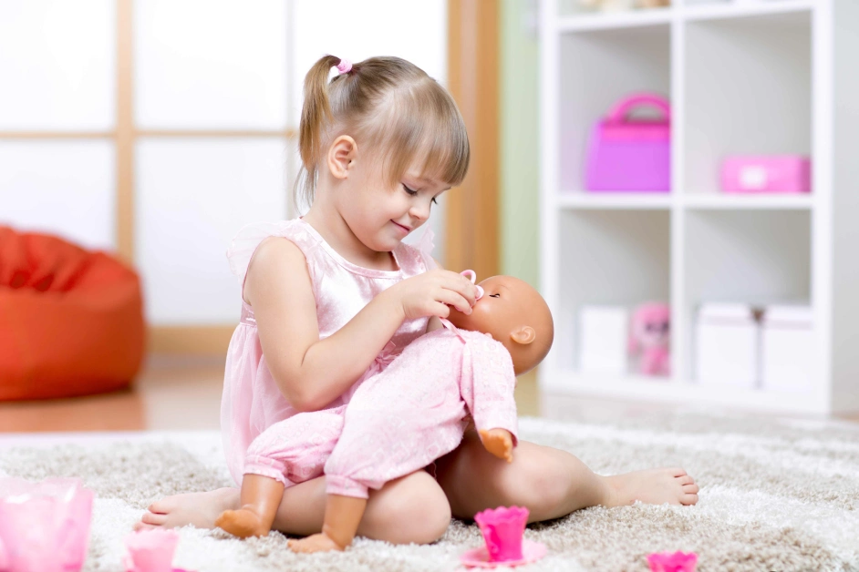 A smiling toddler girl in a pink dress plays with Cute Dolls for Toddlers, gently feeding her baby doll in a cozy, colorful playroom.