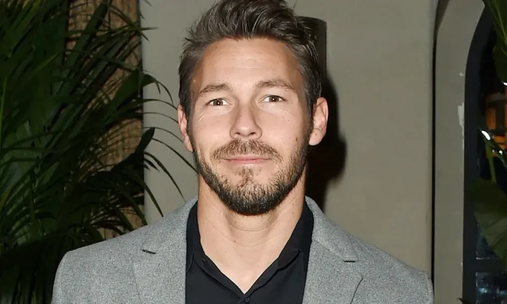 Scott Clifton in 2025 – TV Career, Private Life & Highlights Scott Clifton dressed in a stylish grey blazer and black shirt, standing at an indoor event with lush green plants in the background.