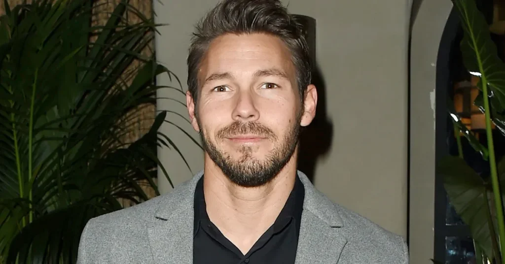 Scott Clifton dressed in a stylish grey blazer and black shirt, standing at an indoor event with lush green plants in the background.