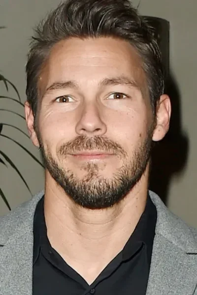 Scott Clifton dressed in a stylish grey blazer and black shirt, standing at an indoor event with lush green plants in the background.